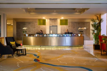 Warm hotel reception with two friendly staff at desk, lounge seating and plants, evening light