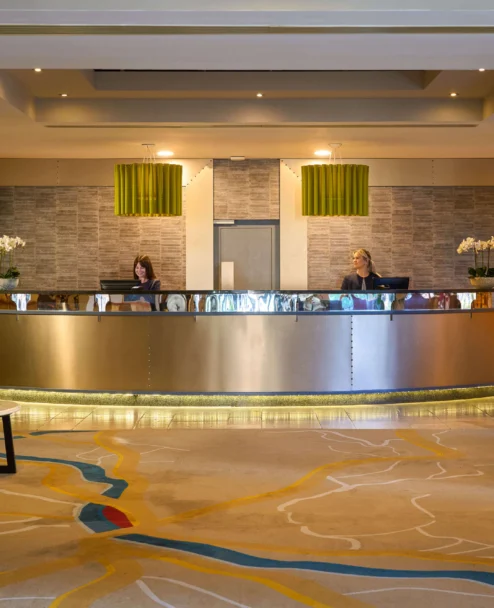 Warm hotel reception with two friendly staff at desk, lounge seating and plants, evening light