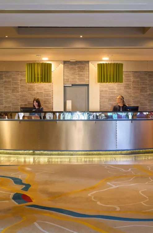 Warm hotel reception with two friendly staff at desk, lounge seating and plants, evening light