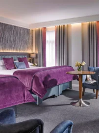 Spacious hotel twin room with plush purple bedding, cosy seating and daytime light through drapes