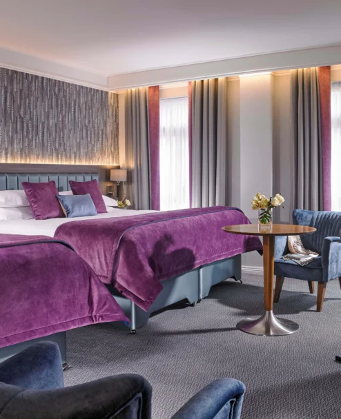 Spacious hotel twin room with plush purple bedding, cosy seating and daytime light through drapes