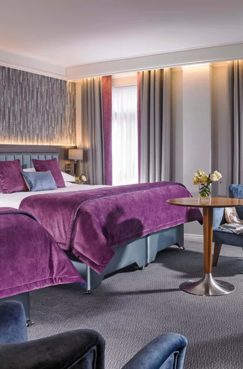 Spacious hotel twin room with plush purple bedding, cosy seating and daytime light through drapes