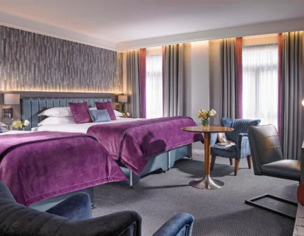Spacious hotel room with two double beds, family-friendly seating and purple linens, warm daylight curtains (no visible text)