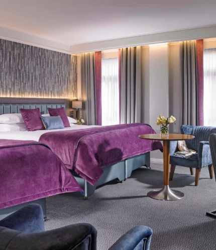 Spacious hotel room with two double beds, family-friendly seating and purple linens, warm daylight curtains (no visible text)
