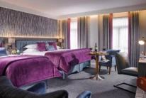 Spacious hotel family room with two beds, purple linens, cosy seating and wine on a table