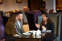 Couple and hotel host smiling over tea in a cosy lounge, warm family-friendly setting (no visible text)