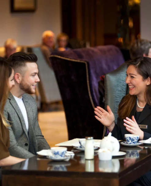 Couple and hotel host smiling over tea in a cosy lounge, warm family-friendly setting (no visible text)
