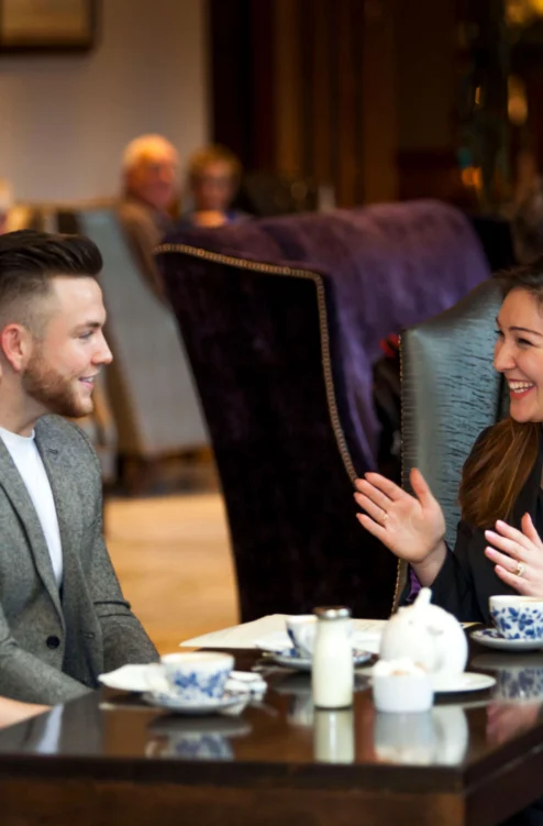 Couple and hotel host smiling over tea in a cosy lounge, warm family-friendly setting (no visible text)