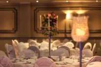 Elegant hotel banquet table with covered chairs, glassware and candlelit floral centrepiece, warm ambience