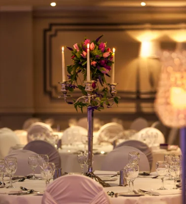 Elegant hotel banquet table with covered chairs, glassware and candlelit floral centrepiece, warm ambience