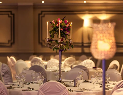 Elegant hotel banquet table with covered chairs, glassware and candlelit floral centrepiece, warm ambience