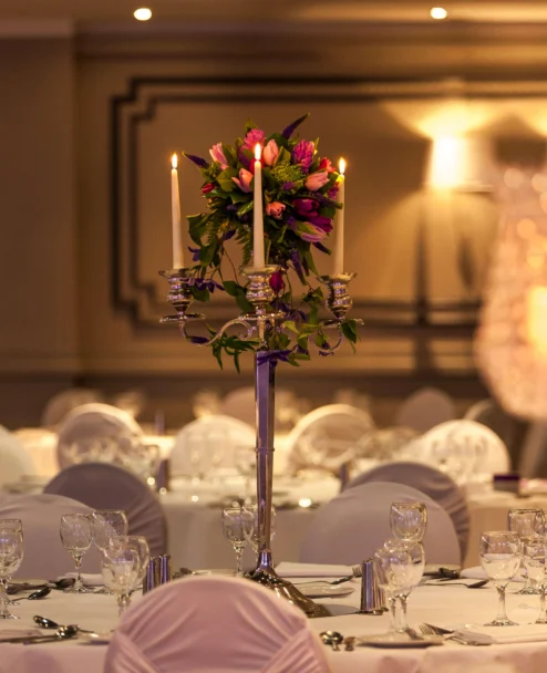 Elegant hotel banquet table with covered chairs, glassware and candlelit floral centrepiece, warm ambience