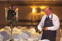 Hotel staff polishing glassware beside a candlelit floral centrepiece as family-style banquet is prepared