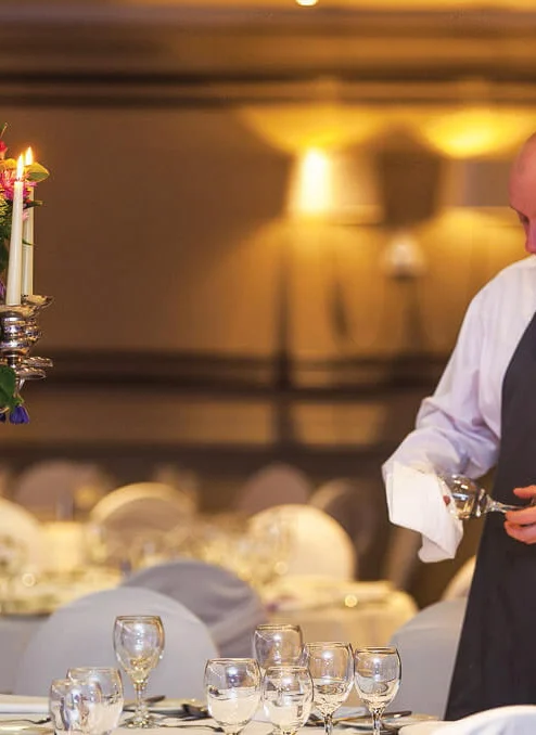 Hotel staff polishing glassware beside a candlelit floral centrepiece as family-style banquet is prepared