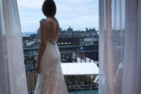 Bride in lace gown on hotel balcony overlooking coastal town at dusk, peaceful moment (no visible text)