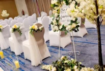 Family wedding setup in a hotel function room with white chair covers, floral aisle decor and candles