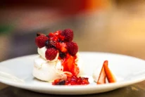 Pavlova dessert topped with fresh raspberries and strawberry sauce on a white plate, close-up, hotel dining