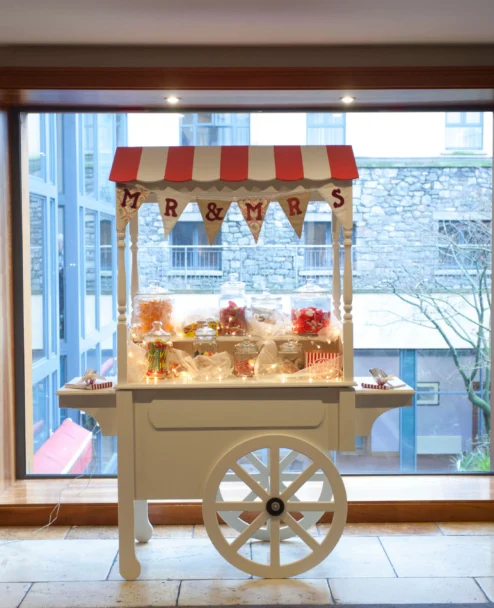 Candy cart with "MR & MRS" bunting and jars of sweets by a hotel window, warm family treat display