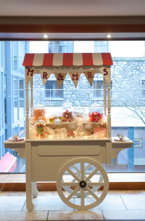 Candy cart with "MR & MRS" bunting and jars of sweets by a hotel window, warm family treat display