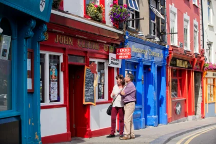 Couple strolling and laughing outside colourful seaside pubs with hanging flowers, shop signs visible