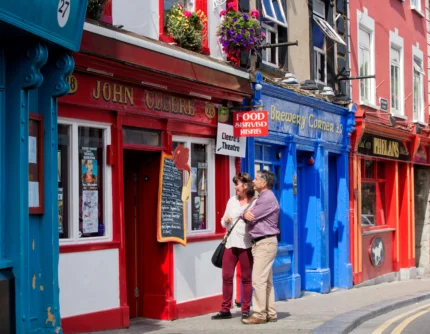 Couple strolling and laughing outside colourful seaside pubs with hanging flowers, shop signs visible