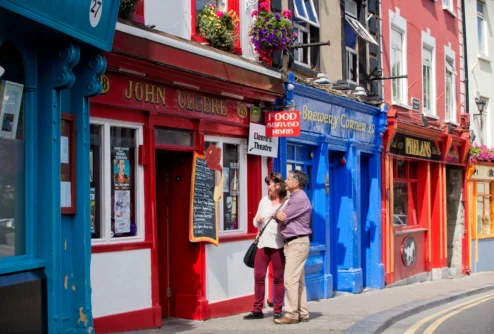 Couple strolling and laughing outside colourful seaside pubs with hanging flowers, shop signs visible
