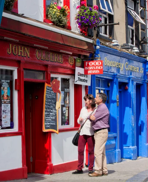 Couple strolling and laughing outside colourful seaside pubs with hanging flowers, shop signs visible