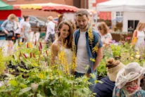Smiling couple browsing colourful flowers at a sunny outdoor market, relaxed family outing, stalls visible