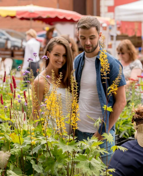 Smiling couple browsing colourful flowers at a sunny outdoor market, relaxed family outing, stalls visible