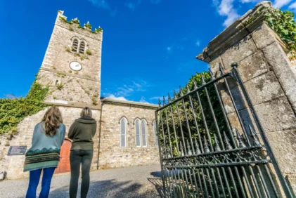 Family exploring a historic stone church courtyard under a bright blue sky, iron gate visible