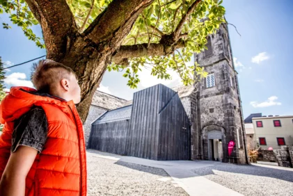 Child in orange jacket with family exploring a sunny town square by historic stone church and trees