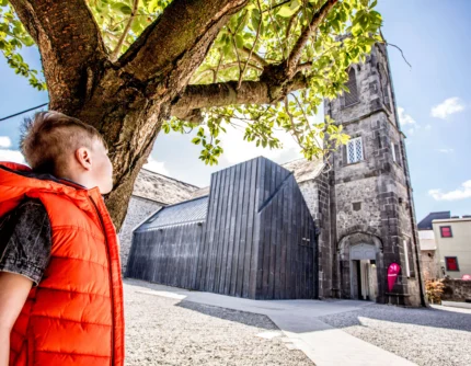 Child in orange jacket with family exploring a sunny town square by historic stone church and trees