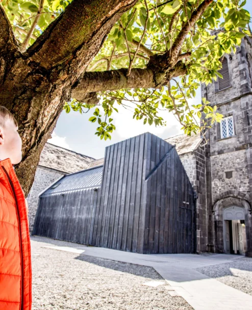 Child in orange jacket with family exploring a sunny town square by historic stone church and trees