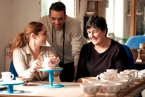 Family of three smiling together over tea and hand‑painted pottery in a cosy studio (no visible text)