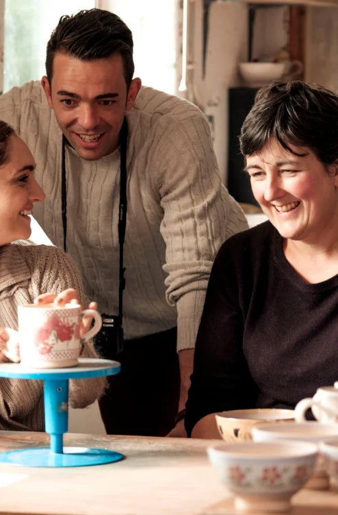 Family of three smiling together over tea and hand‑painted pottery in a cosy studio (no visible text)
