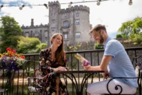 Smiling couple on a sunny terrace by a historic castle, enjoying a brochure and relaxed conversation
