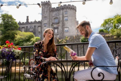 Smiling couple on a sunny terrace by a historic castle, enjoying a brochure and relaxed conversation