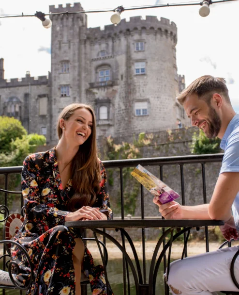 Smiling couple on a sunny terrace by a historic castle, enjoying a brochure and relaxed conversation