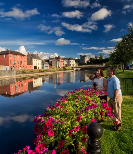 Couple enjoying riverside stroll by colourful townhouses and a castle across calm water, summer sky