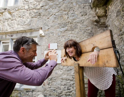 Smiling couple playfully posing in wooden stocks against a stone courtyard wall at a historic hotel attraction