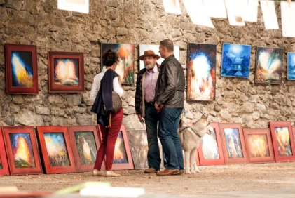 Family browsing colourful framed art at an outdoor market against a stone wall, dog at their feet