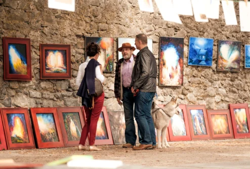 Family browsing colourful framed art at an outdoor market against a stone wall, dog at their feet