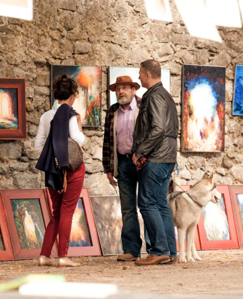 Family browsing colourful framed art at an outdoor market against a stone wall, dog at their feet