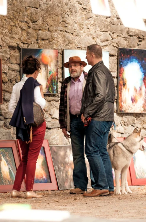 Family browsing colourful framed art at an outdoor market against a stone wall, dog at their feet