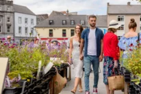 Couple holding hands strolling past colourful market flowers in a sunny town square, family atmosphere