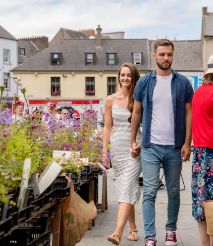 Couple holding hands strolling past colourful market flowers in a sunny town square, family atmosphere