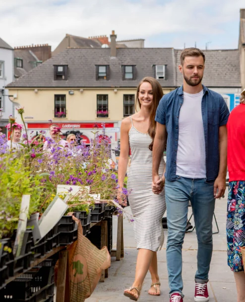 Couple holding hands strolling past colourful market flowers in a sunny town square, family atmosphere
