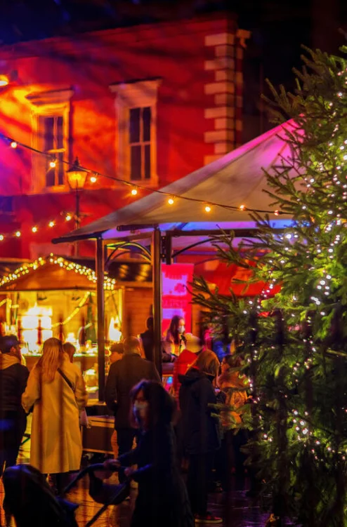 Family exploring a festive night market by lit stalls and a decorated Christmas tree, cosy atmosphere