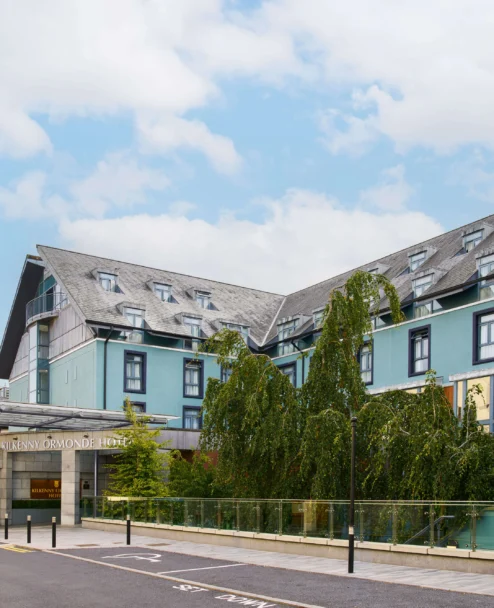 Family checking into a welcoming seaside hotel entrance with blue façade and leafy trees, sign reads "KILKENNY ORMONDE HOTEL" (70–100 chars)
