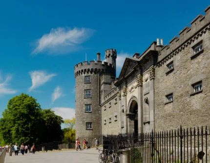 Family strolling near historic stone castle entrance with blue sky, trees and bicycles